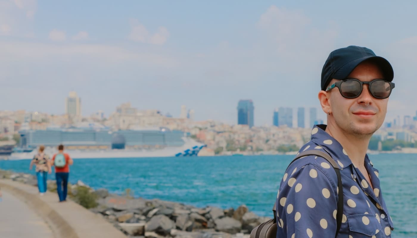 Man wearing a navy cap, sunglasses, and a polka-dot shirt stands on a waterfront promenade with a city skyline and a cruise ship in the background.